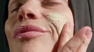 Kapalı mekanda, tesettürlü kadın yeşil cilt ürününü nazikçe uyguluyor yanağı kendi kendine bakım rutinine odaklanıyor. Işık dingin bir atmosfer yaratır.