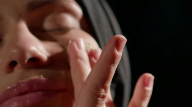 Başörtüsü takan kadın, sıcak evinde, gece bakım rutininin bir parçası olarak nazikçe cilt bakım ürününü uyguluyor. Ilık aydınlatma sakinleştirici atmosfer yaratır.