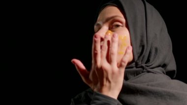 Siyah tesettürlü bir kadın, loş aydınlatılmış odada kültürel güzellik uygulamaları sergileyerek yüzüne canlı sarı kozmetik tedavi uyguluyor..