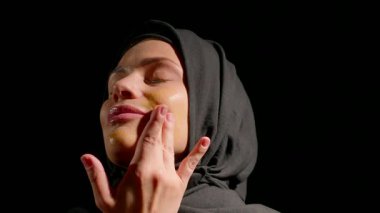 Siyah tesettürlü bir kadın doğal cilt bakımı uygular. Yüzü huzur ve rahatlığı yansıtır. Sıcak ışık odaklanmış ifadesini vurguluyor..