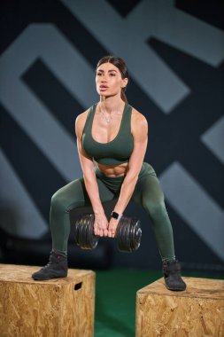 Kadın spor salonunda dambılları tutarken çömelme egzersizi yapıyor. Güç antrenmanı rutini üzerinde çalışırken odaklanmış ve kararlı..