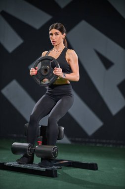Aktif giysili bir kadın spor salonunda antrenman yapıyor. Ağırlık levhasını tutarken bankta oturur ve egzersiz rutinine odaklandığını gösterir..