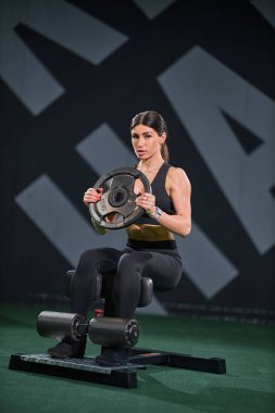 Kendini işine adamış bir kadın spor salonunda ağırlık plakası kullanarak egzersiz yapmaya odaklanmış. özelliklerin ayarlanması spor aletleri ve motivasyonel duvar grafikleri.