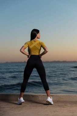 Kadın gün batımında sarı bluz ve siyah tayt giyerek güvenle ayakta duruyor. Sakinleştirici dalgalar ve yumuşak ışık o güç yaydıkça sakin bir atmosfer yaratır..