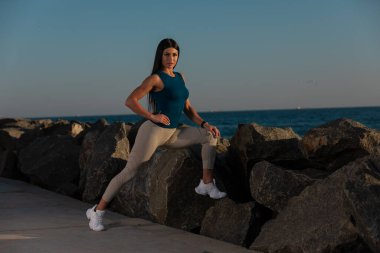 Kadın, arka planda güneş batarken kayalık kıyı şeridinde esneme egzersizleri yapıyor. Okyanus dalgaları, o egzersizine odaklanırken sakin bir atmosfer yaratır..