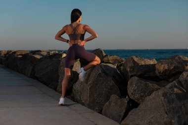 Kadın sırtını kayalık yolda geriyor. Spor giysileri giyiyor ve egzersiz rutinine hazırlanırken huzurlu bir gün batımının keyfini çıkarıyor..