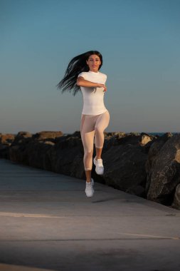 Hafif giyimli bir kadın sakin sahil yolu boyunca koşar. Gün batımı kararlılığını gösterir ve zemin güzelliğinin arasında zindelik ve sağlığa odaklanır..