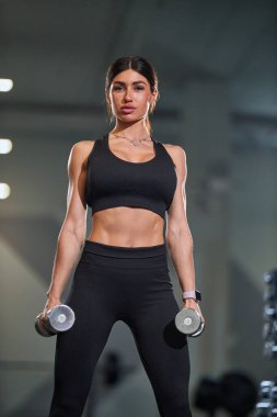 Fitness meraklısı modern spor salonunda dambılları kaldırarak dayanıklılığını artırıyor. Atmosfer, arka plandaki spor aletlerini motive ediyor ve çalıştırıyor.