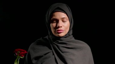 Siyah tesettürlü bir kadın sessizce dikiliyor. Gözleri kapalı. Elinde, meditasyona daldırılmış kırmızı bir gül var..