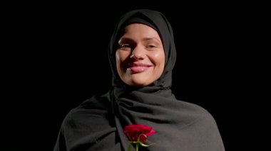 Siyah başörtüsü takan kadın, kırmızı gülü tutarken kendine güvenerek gülümsüyor ve kokluyor. Arka plan koyu renkli, neşeli ifadesini ve canlı çiçeğini vurguluyor..