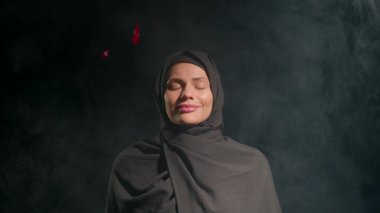 Siyah tesettürlü bir kadın, sessiz uzayda gözlerini kapadığı yerde hareketsiz duruyor. Yumuşak duman onu sararken kırmızı taçyapraklar sükunet veren atmosferin üzerinde yavaşça süzülüyor..