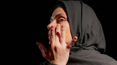Siyah tesettürlü kadın nazikçe cilt kremi sürüyor. Yüzü, parmakları, hafifçe aydınlatılmış alanda odaklanma ve ilgi gösteriyor. Huzurlu bir atmosfer yaratıyor..