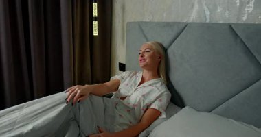 Kız yatakta kulaklıkla rahatça oturuyor ve telefonda konuşurken gülümsüyor. Güneş ışığı perdelerin arasından yavaşça süzülür ve evde rahat bir atmosfer yaratır..