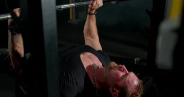 Adam ağırlık kaldırma egzersizlerinde ağırlık kaldırmaya odaklanmış. Kulaklık takıyor ve iyi donanımlı bir spor salonunda eğitim görüyor..