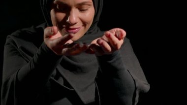 Siyah tesettüre bürünmüş kadın, elinde nazikçe bir nesne tutarken sevinçten havalara uçuyor. Sıcak gülüşü, yansıma anını vurgulayan koyu arkaplanı zıtlaştırıyor..