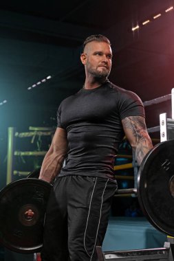 Geniş bir spor salonunda halter egzersizleri yapan güçlü bir adam. Atmosfer enerjiktir. Parlak ışıklar onun kararlı ifadesini ve kaslı fiziğini vurgular..