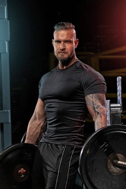 Kaslı adam spor salonunda halter egzersizi yapıyor. Her elinde bir halter tutarken yoğun bir konsantrasyon sergiliyor. Her iki elinde de zindelik ve dayanıklılık gösteriyor..
