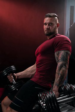 Kaslı bir adam loş bir spor salonunda ağır dambılları kaldırarak antrenman yapıyor. Ortam egzersizler için odaklanmış bir atmosfer yaratıyor..
