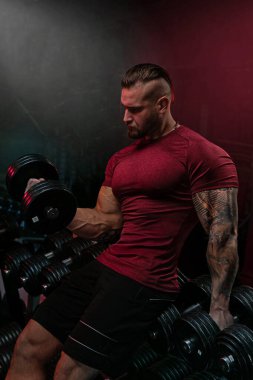 Kırmızı gömlekli kaslı adam spor salonunda dambıl kaldırıyor. Etrafı çeşitli ağırlıklarla çevriliyken odaklanmış ve kararlı görünüyor. Işıklandırma yoğun bir atmosfer yaratır..