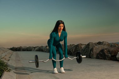 Deniz mavisi spor kıyafetli genç bir kadın güneş batarken kayalık sahilin yanındaki halteri kaldırıyor. Sahil sporu dingin ama güçlendirici spor anını yakalar..