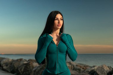 Model kumsalda duruyor, atletik giysiler içinde sakin okyanus dalgalarının ve gün batımında renkli gökyüzünün ortasındaki mesafeye bakıyor..