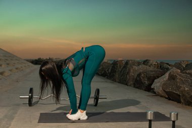 Antrenman kıyafetli kişi kayalık kıyı şeridi boyunca yer alan minder üzerinde öne eğilme hareketi yapıyor. Canlı günbatımı gökyüzünü aydınlatıyor sıcak renkler sakin bir atmosfer yaratıyor.
