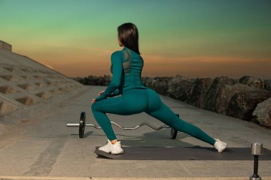 Deniz mavisi giysili kadın spor yapıyor halterler üzerinde dans ediyor. Gün batımı renkli bir zemin oluşturur. Antrenman yaptığı kayalık kıyı şeridini güçlendirir..