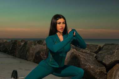 Fitness modeli, kıyı yolunda dinamik egzersiz pozuyla meşguldür. Etrafı kayalar ve sakin okyanuslarla çevrilidir. Gün batımı canlı bir zemin oluşturur zenginleştirici sahne.