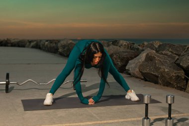 Ağırlıklar yakınlarda otururken, bireysel uygulamalar deniz kenarındaki mindere uzanır. Güneş arka planda batar ve egzersiz için sakin bir atmosfer yaratır.