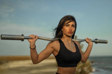 Kararlı kadın sahilde omuzlarının üzerinden halter kaldırıyor. Güneş batıyor, sahneye sıcak bir parıltı saçıyor. Gücünü ve odaklanmasını vurguluyor..