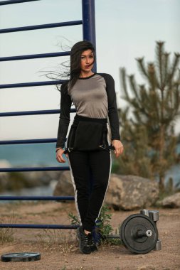 Genç bir kadın açık hava ortamında bir spor aletinin yanında duruyor. Günlük spor kıyafetleri giyer ve güneşin ve doğanın tadını çıkarırken kendinden emin bir poz verir..