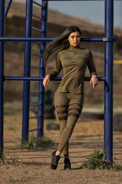 Genç bir kadın spor kıyafetleri içinde zarif bir şekilde oyun sahasında duruyor. O kendine güven yayarken güneş ışıl ışıl parlıyor. Arka planda fitness ekipmanları var.