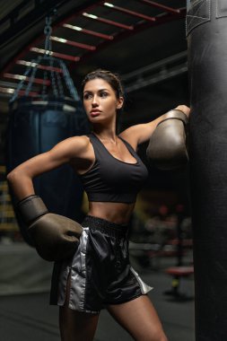 Boks eldiveni ve şort giyen kadın boksör iyi aydınlatılmış bir spor salonunda ağır bir kum torbasına vurmaya hazırlanıyor. Sahne, akşam antrenmanı sırasında dikkatini ve gücünü çekiyor..