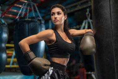 Yetenekli bayan boksör bir spor salonunda gücünü ve kararlılığını gösteriyor. Eldiven takıyor ve spor kıyafeti giyiyor. Ağır bir kum torbasının yanında güçlü bir poz veriyor..