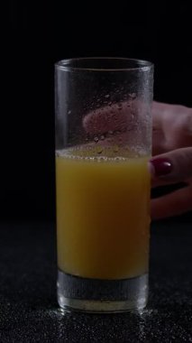 Kadınların elleri nazikçe taze portakal suyuyla dolu soğuk bir bardağa uzanır. Arkaplan, ferahlatıcı içkiyi vurgulamak için karanlık. Camdaki buğulanma soğuğu vurguluyor..