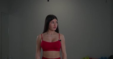 Sportif kız, spor salonundaki spor malzemeleriyle dolu spor salonunda kameraya kendinden emin bir ifadeyle bakıyor..