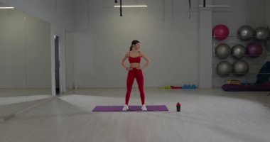 Parlak bir spor salonundayken kız boynunu esnetiyor. Çevreleme ekipmanları aktif bir çevreyi gösteren ağırlık ve egzersiz toplarını içerir.