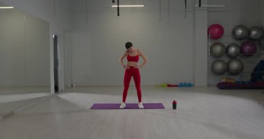 Geniş bir spor salonunda kırmızı spor kıyafetli bir kız paspasın üzerinde dikilirken boynunu uzatıyor. Etkin bir çalışma oturumunu gösteren egzersiz ekipmanları yakınlarda düzenlenmiştir.