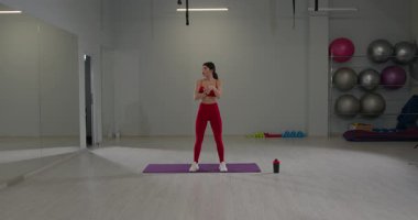 Kız, spor salonundaki yoga minderinde el yoğurma egzersizi yapıyor. Stüdyoda aynalar ve etkin bir ortam yaratmak için çeşitli çalışma ekipmanları bulunuyor..
