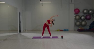 Geniş bir spor salonunda spor kıyafeti giyen bir kız yoga minderinde egzersiz yaparken yana doğru eğilir..