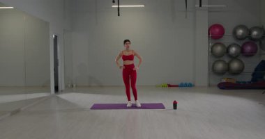 Kırmızı giysili kız, parlak ferah bir spor salonunda antrenman yapmaya hazırlanıyor. Odada egzersiz ekipmanları ve renkli spor aksesuarları bulunuyor..