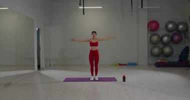 Kız, arka planda egzersiz ekipmanları olan geniş bir spor salonunda spor kıyafetleri içinde zıplama hareketleriyle meşgul. Atmosfer, enerjik bir egzersizi yansıtıyor..