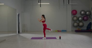 Modern bir fitness stüdyosunda bir kız spor yapar ve mor bir paspasın üzerinde çömelme ve bacak kaldırma hareketleri yapar. Parlak alan spor aletleri ve ağırlıklarla donatılmıştır..
