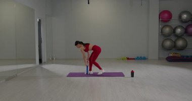 Parlak bir fitness stüdyosunda aktif giyimli bir kız bel egzersizi yapmak için lastiği çeker. Odaklanması ve biçimi egzersizler sırasında güç eğitiminin önemini vurguluyor..