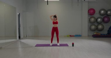 Geniş bir spor salonunda, spor kıyafeti giymiş bir kız mor bir paspasın üzerinde dururken omuzlarını ve trisepslerini uzatır. Yakınlarda bir su şişesi fitness takımını tamamlıyor..