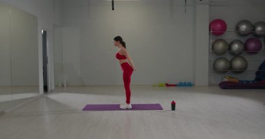 Kırmızı spor kıyafetli bir kız spor salonunda bacaklarını ve sırtını esnetiyor. Çevrede egzersiz ekipmanları ve yerde bir yoga minderi bulunuyor..