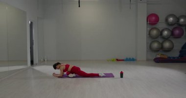 Kız geniş bir spor salonundaki mor bir paspasın üzerinde esneme hareketleri yapıyor. Esnekliği artırmak için sırtına ve bacaklarına odaklanıyor..