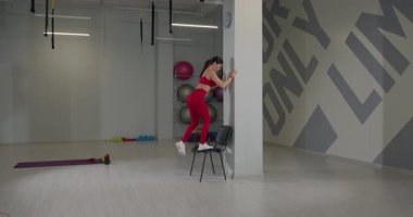 Kırmızı giysili kadın, parlak bir spor salonundaki sandalyeye basıyor. Zeminde düzenli olarak çeşitli egzersiz ekipmanları düzenlenmiştir. Canlı bir egzersiz atmosferi sergilerler..