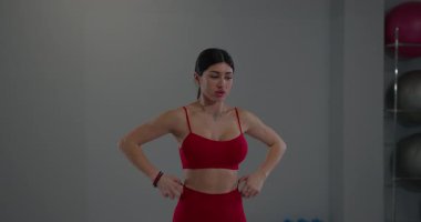 Kız parlak kırmızı bir spor kıyafeti giyerken boynunu esnetiyor. Hareketine ve duruşuna odaklanmış egzersiz ekipmanlarıyla çevrili iyi aydınlatılmış bir spor salonunda..