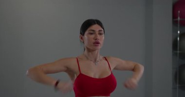 Modern bir spor salonunda omuz germe hareketleri yapan bir kız. Spor aletleri ve rahatlatıcı bir atmosferle çevrili kırmızı bir bluz giyerek formuna odaklanıyor..
